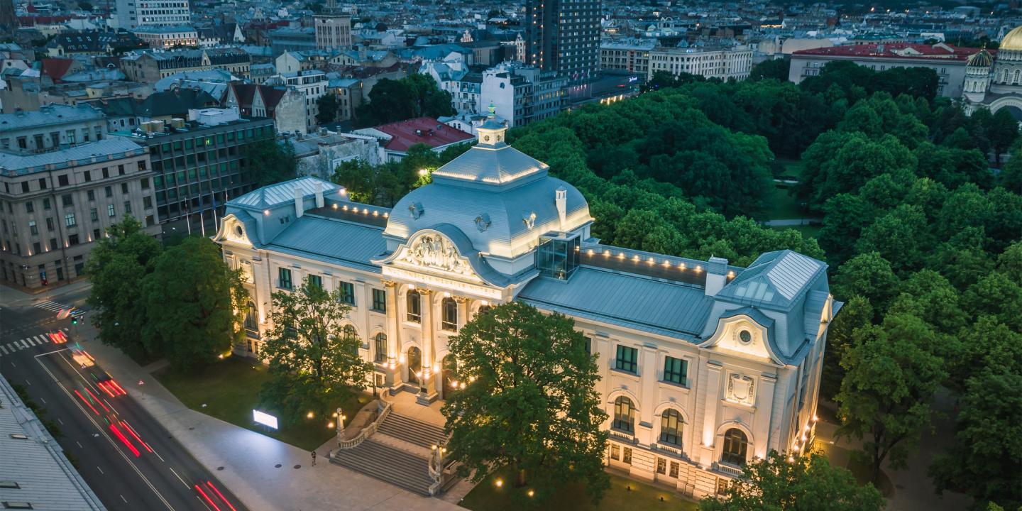 Apgaismota Latvijas Nacionālā mākslas muzeja ēka Rīgā krēslā, skatīta no augšas ar pilsētas panorāmu fonā.