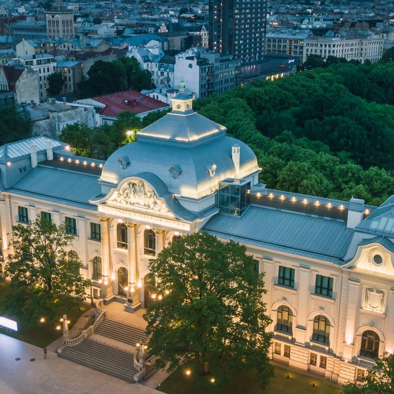 Apgaismota Latvijas Nacionālā mākslas muzeja ēka Rīgā krēslā, skatīta no augšas ar pilsētas panorāmu fonā.
