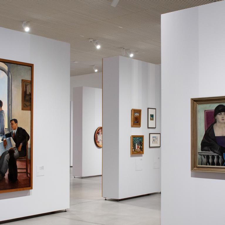 Muzeja izstāžu zāle ar gleznām pie baltām sienām, centrā redzamas figurālas kompozīcijas un portreti.