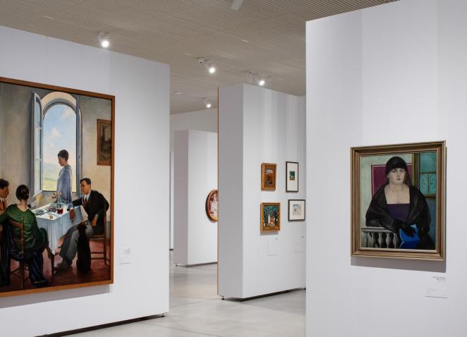 Muzeja izstāžu zāle ar gleznām pie baltām sienām, centrā redzamas figurālas kompozīcijas un portreti.