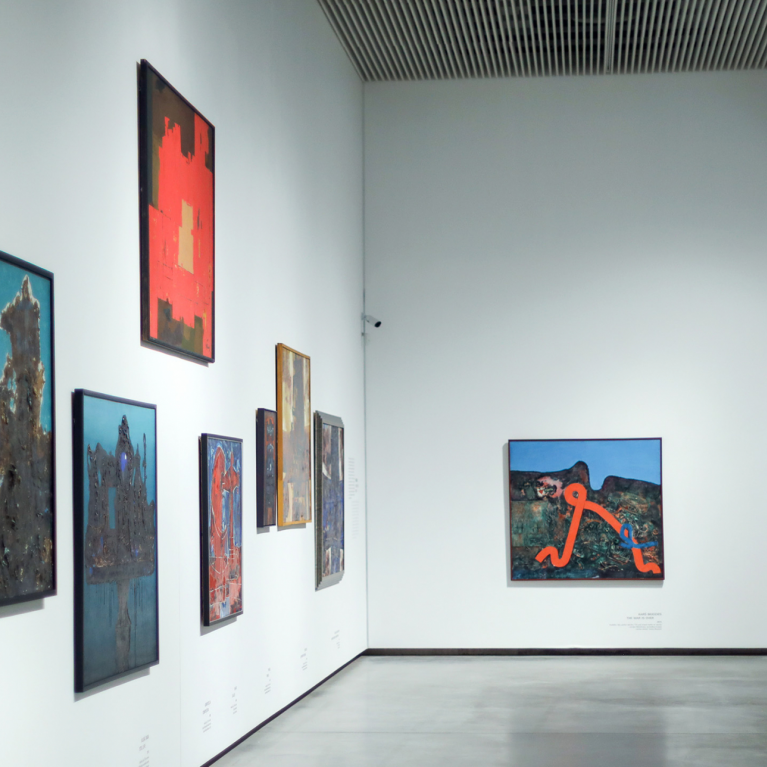 Museum exhibition hall with white walls displaying multiple paintings in various formats; a spacious, minimalist interior view.