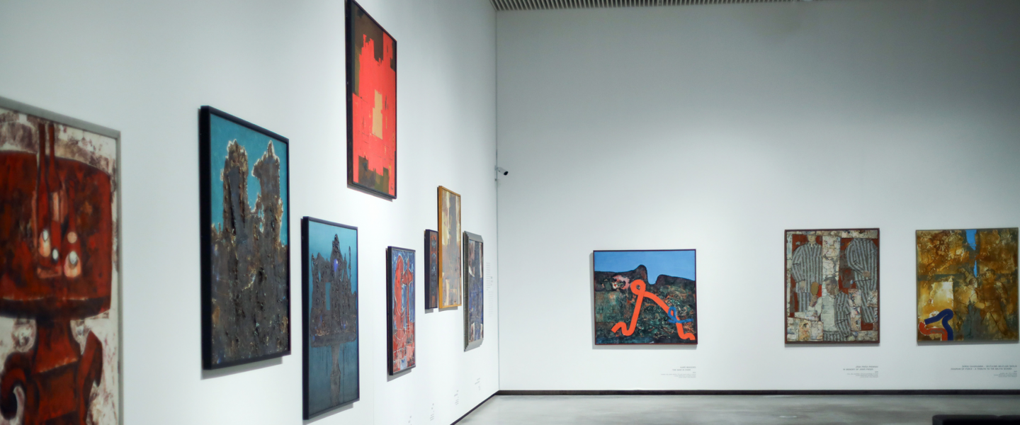 Museum exhibition hall with white walls displaying multiple paintings in various formats; a spacious, minimalist interior view.