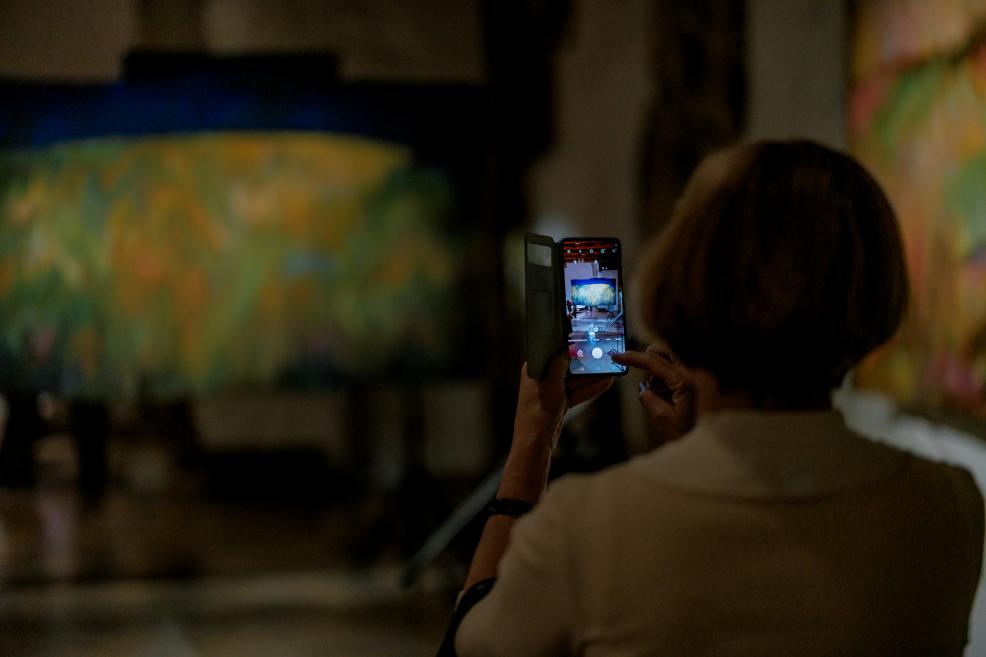 Sieviete izstādē fotografē krāsainu mākslas darbu ar savu telefonu.