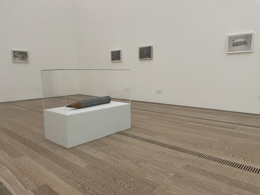 A minimalist exhibition space with a glass display case at the center showcasing an enlarged pencil-shaped object, surrounded by small graphic works on white walls, creating a quiet and contemplative viewing atmosphere.