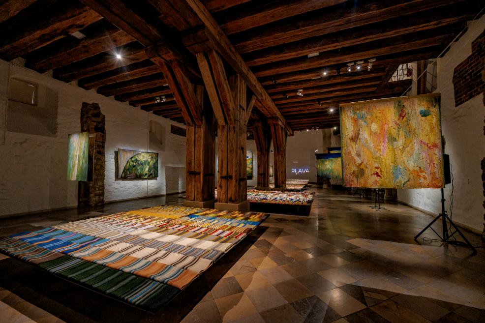 An exhibition hall with wooden beams, displaying multi-colored tapestries on the floor and walls.