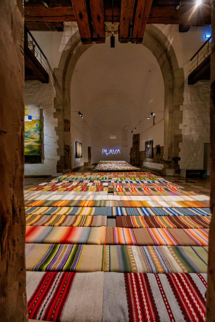 An exhibition hall with colorful striped textiles on the floor and tapestries on the walls; the word 'PLAVA' shines in the background.