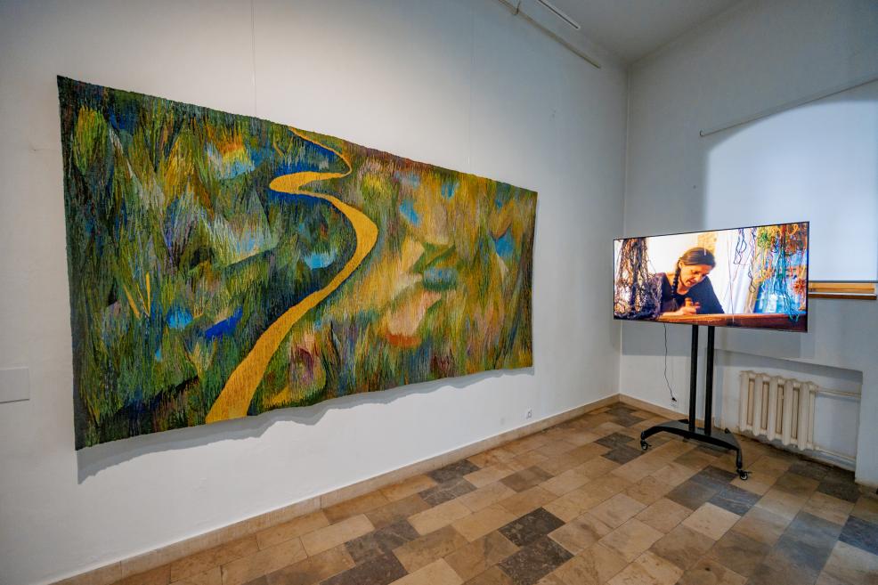 An exhibition corner with a tapestry depicting a yellowish road in a landscape, and a screen next to it with a video of the artist in action.
