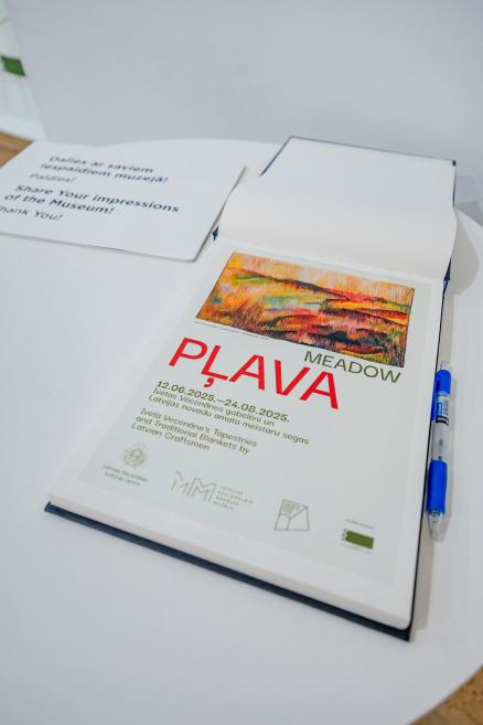 Atsauksmju grāmata ar izstādes 'Pļava / Meadow' afišu uz vāka un pildspalvu uz galda.
