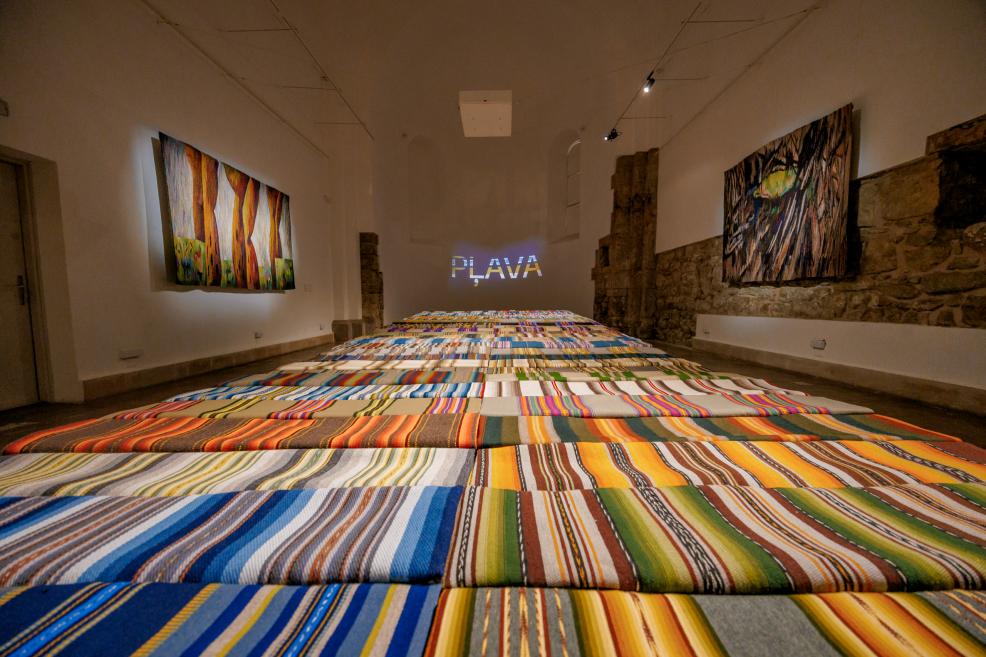 An exhibition hall with colorful striped textiles on the floor and tapestries on the walls; the word 'PLAVA' shines in the background.
