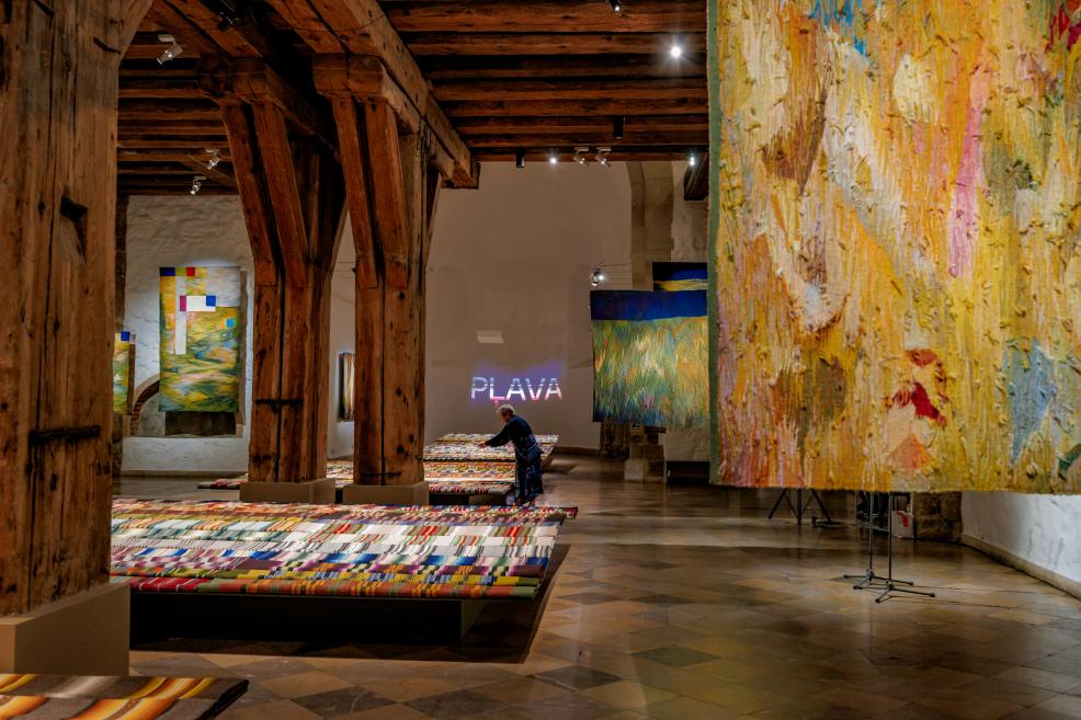 The 'Meadow' exhibition hall with wooden pillars, colorful tapestries on the walls, and textiles on the floor; a visitor and the illuminated inscription 'Meadow' in the background.