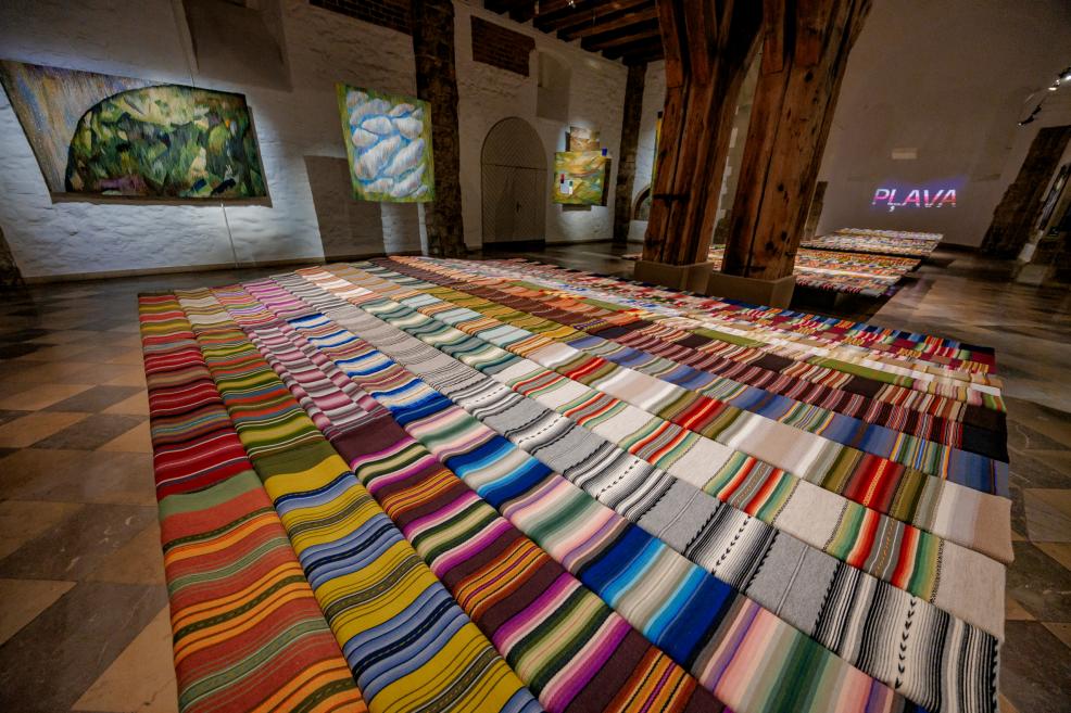 The 'Pļava' exhibition room with bright striped textiles on the floor and tapestries on the walls; the inscription 'PLAVA' is illuminated in the background.