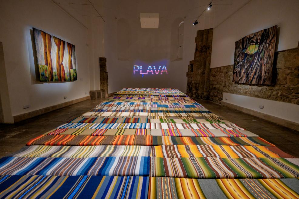 The 'Meadow' exhibition hall with multi-colored fabrics on the floor and tapestries on the walls; the exhibition title 'PLAVA' is illuminated in the background.