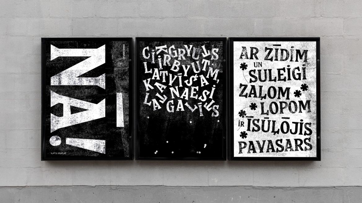 Uz pelēkas sienas blakus izkārtoti trīs melnbalti tipogrāfiski plakāti melnos rāmjos: kreisajā liels stilizēts burts, vidū izkaisīti burti, labajā latvisks teksts ar lieliem vārdiem.