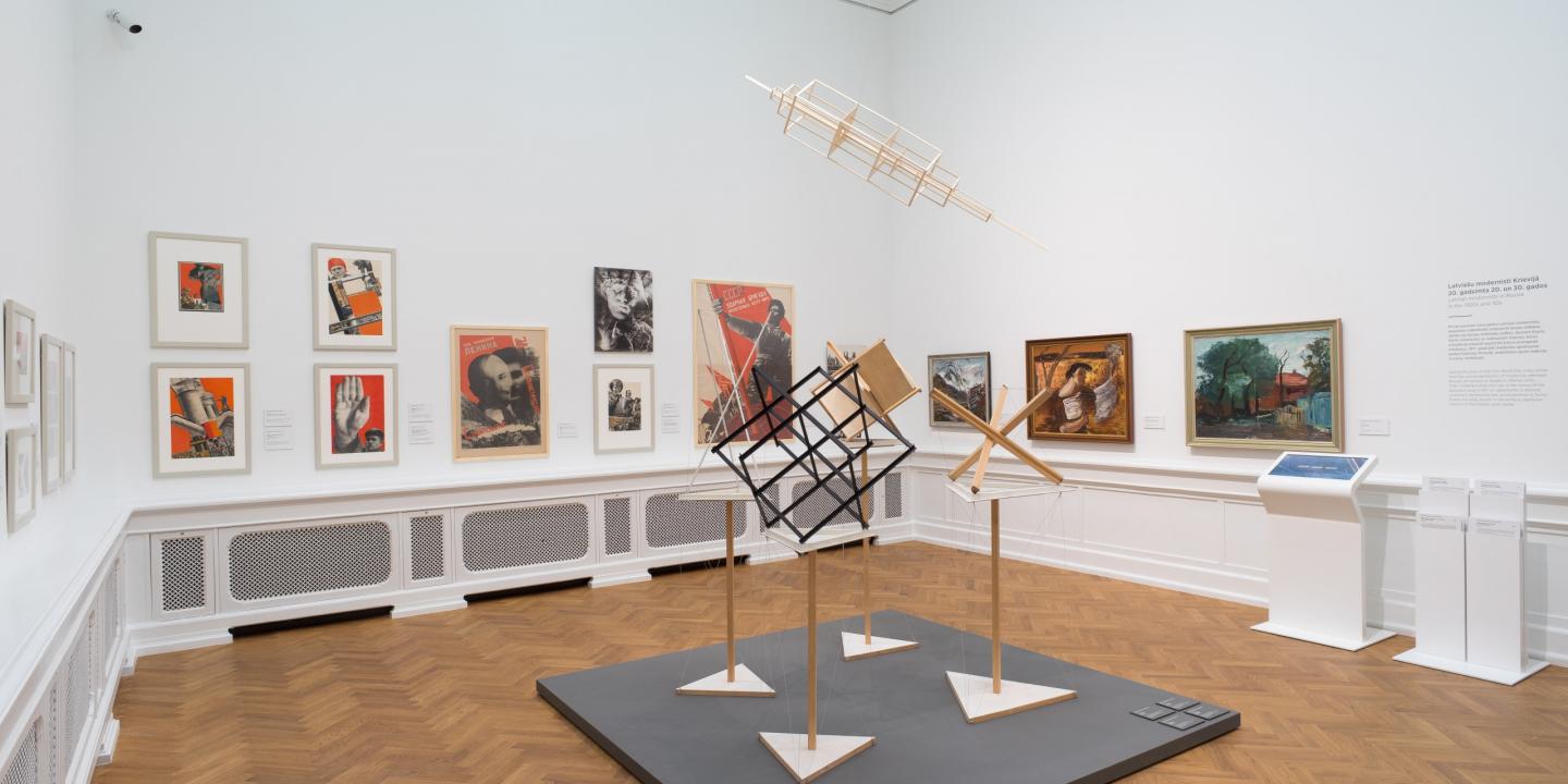 Izstādes zāle ar 20. gadsimta mākslas darbiem — gleznām, fotomontāžām un konstruktīvisma iedvesmotām telpiskām instalācijām, eksponētām uz baltām sienām un koka grīdas fona.