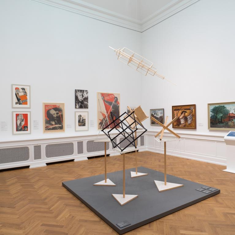 Izstādes zāle ar 20. gadsimta mākslas darbiem — gleznām, fotomontāžām un konstruktīvisma iedvesmotām telpiskām instalācijām, eksponētām uz baltām sienām un koka grīdas fona.