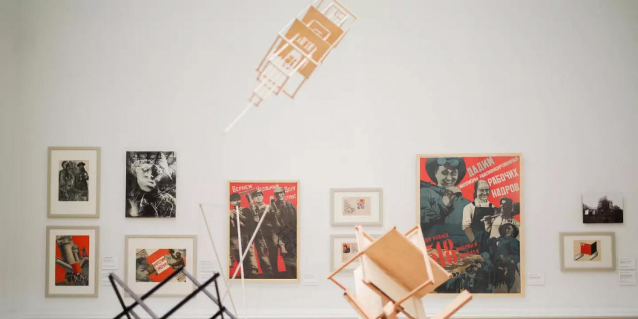 Skats uz izstādes telpu ar 20. gadsimta plakātiem, fotomontāžām un koka konstrukciju instalācijām zāles centrā.