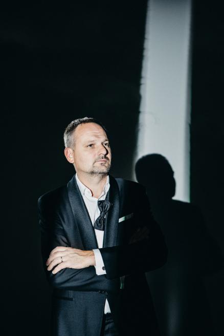 Vīrietis elegantā tumšā uzvalkā stāv pie tumšas sienas ar rokām sakrustotām un skatās sāņus, gaismā veidojot asu ēnu.