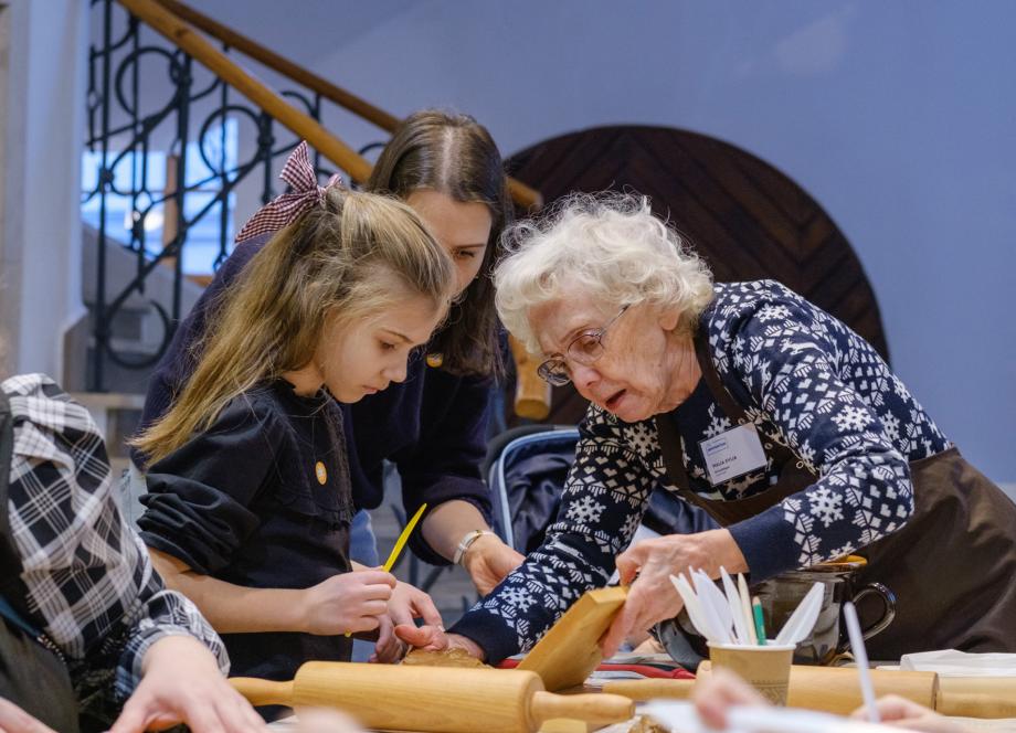 Vecāka sieviete palīdz bērnam veidot roku darbu muzeja nodarbībā, viņus iedrošinot un skaidrojot procesu.