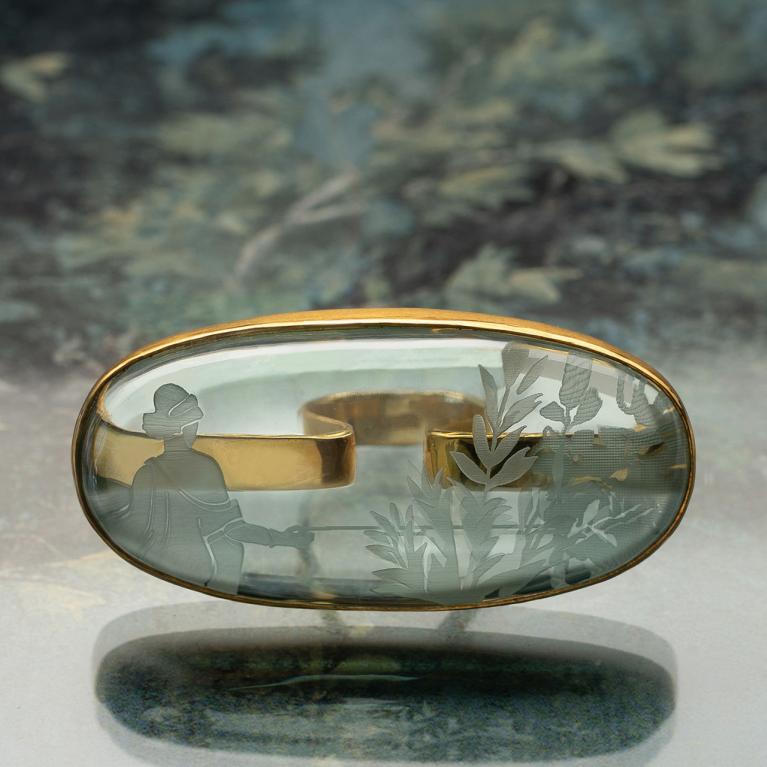 Oval glass ring engraved with plant and human motifs, placed against a softly blurred natural background.