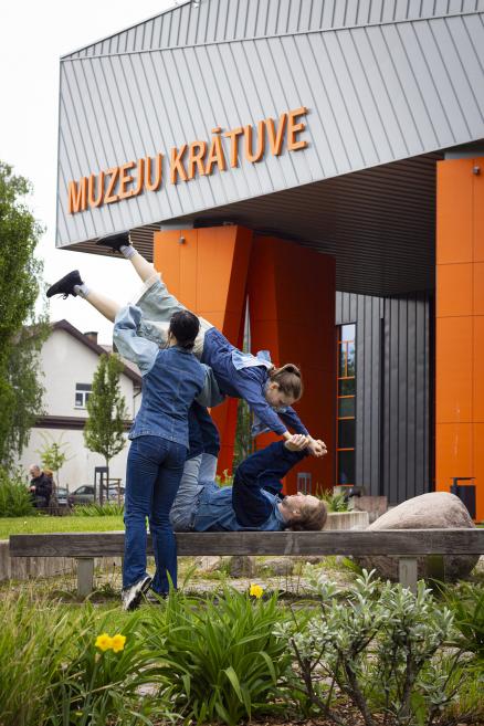 Vairāki cilvēki izpilda kustību performanci pie Muzeju krātuves ēkas, veidojot līdzsvara un pacelšanas pozu uz soliņa ārpusē.