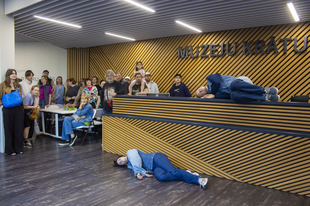 Apmeklētāji vēro performanci Muzeju krātuves vestibilā, kur divi cilvēki atrodas neparastās pozās uz grīdas un pie informācijas letes.