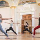 Four women perform Warrior II Pose on yoga mats in a historic museum hall, extending their arms horizontally and maintaining a focused gaze.