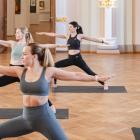 Four women perform Warrior II Pose on yoga mats in a historic museum hall, extending their arms horizontally and maintaining a focused gaze.