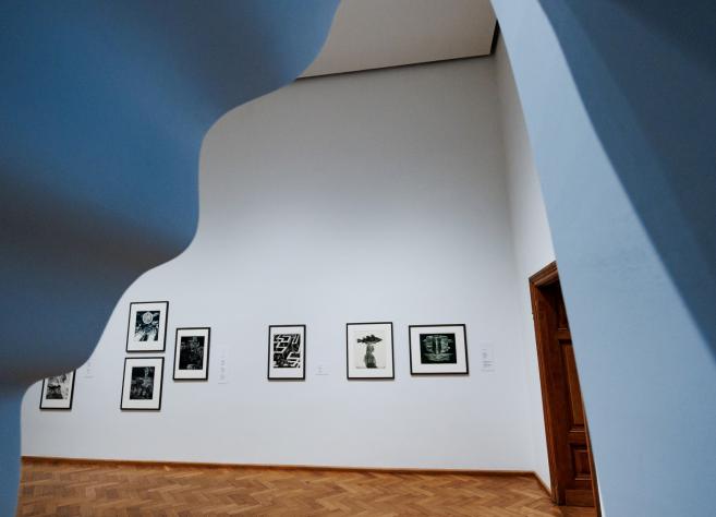 Muzeja ekspozīcijas zāle ar melnbaltām grafikām pie baltas sienas, skatījums caur izliektu arhitektūras atvērumu.