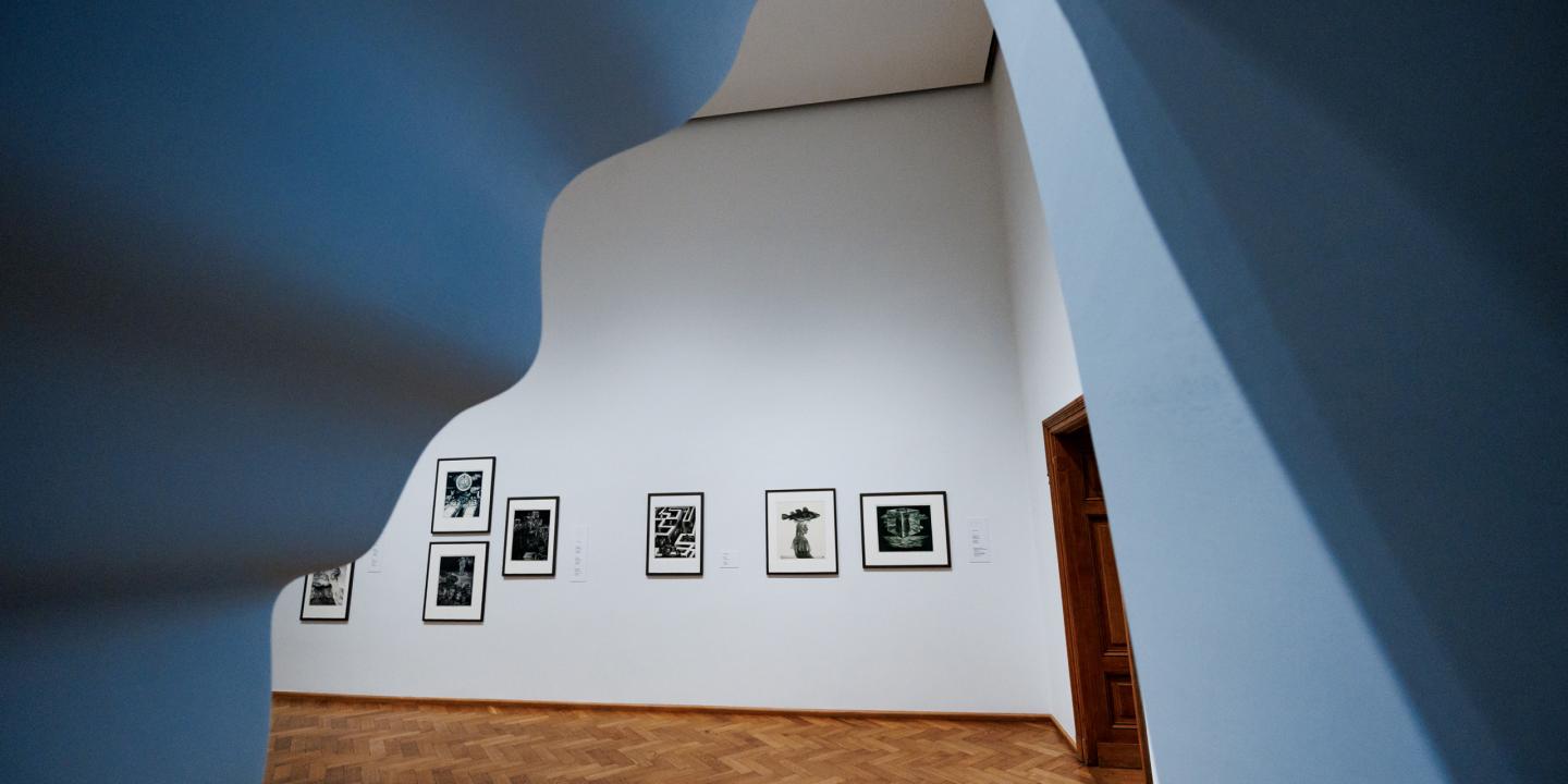 Muzeja ekspozīcijas zāle ar melnbaltām grafikām pie baltas sienas, skatījums caur izliektu arhitektūras atvērumu.