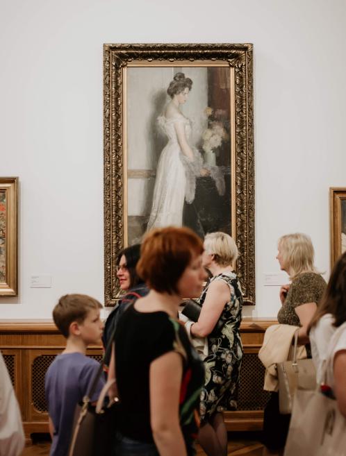 Muzeja zālē pie sienas karājas liela portretglezna zeltītā rāmī, bet priekšplānā stāv apmeklētāji.