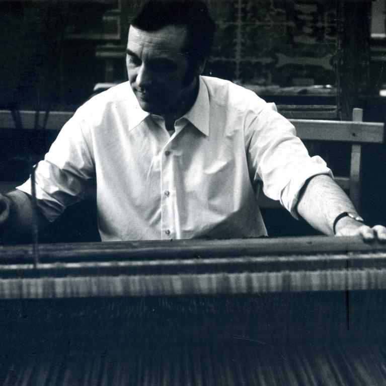 Black-and-white photograph of a man in a white shirt working at a loom or weaving machine.