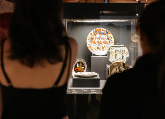 Muzeja izstādē stikla vitrīnā izvietoti vairāki dekoratīvi šķīvji ar ģeometriskiem un figūru motīviem, ko aplūko apmeklētāji.