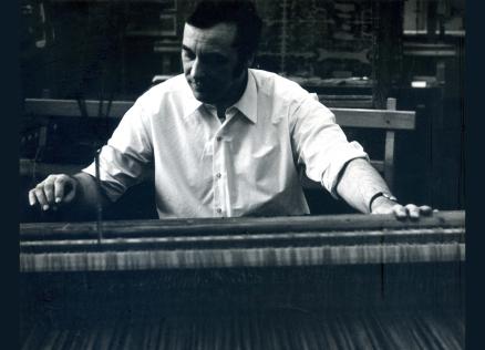 Black-and-white photograph of a man in a white shirt working at a loom or weaving machine.