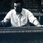 Black-and-white photograph of a man in a white shirt working at a loom or weaving machine.