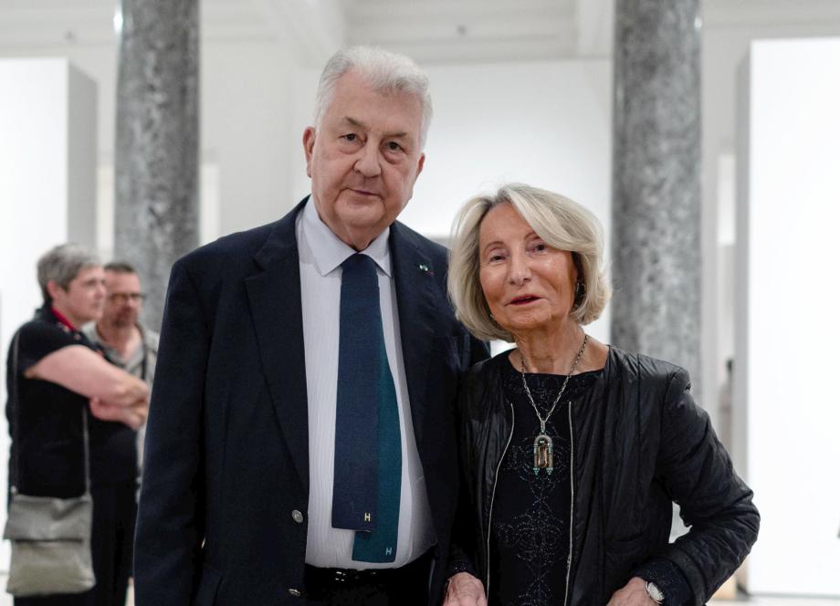Muzeja zālē stāv eleganti ģērbts sirmgalvis uzvalkā un sieviete melnā jakā ar kaklarotu, fonā redzami citi apmeklētāji un baltas ekspozīcijas sienas.