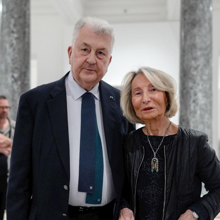 Muzeja zālē stāv eleganti ģērbts sirmgalvis uzvalkā un sieviete melnā jakā ar kaklarotu, fonā redzami citi apmeklētāji un baltas ekspozīcijas sienas.