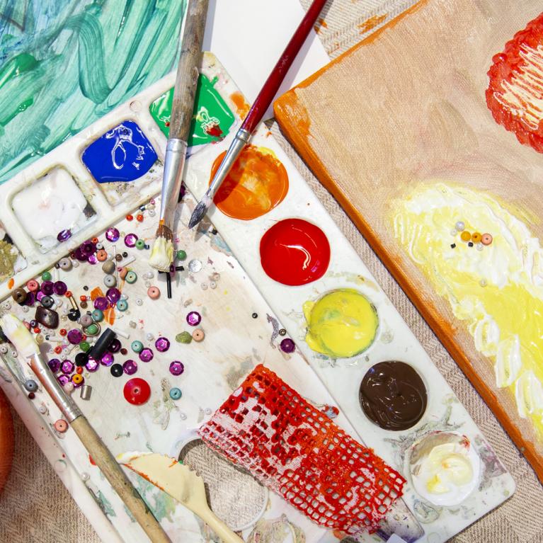 Mākslinieka darba virsma no augšas: krāsu palete ar košām krāsām, otas, krelles un dažādi fakturēti materiāli, blakus audekls ar biezu krāsu klājumu dzeltenos, oranžos un sarkanos toņos.