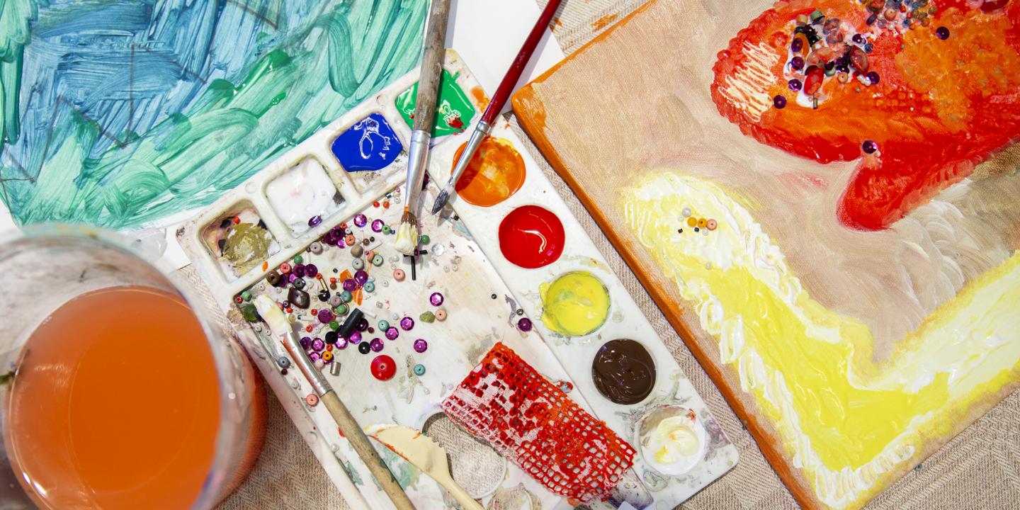 Mākslinieka darba virsma no augšas: krāsu palete ar košām krāsām, otas, krelles un dažādi fakturēti materiāli, blakus audekls ar biezu krāsu klājumu dzeltenos, oranžos un sarkanos toņos.