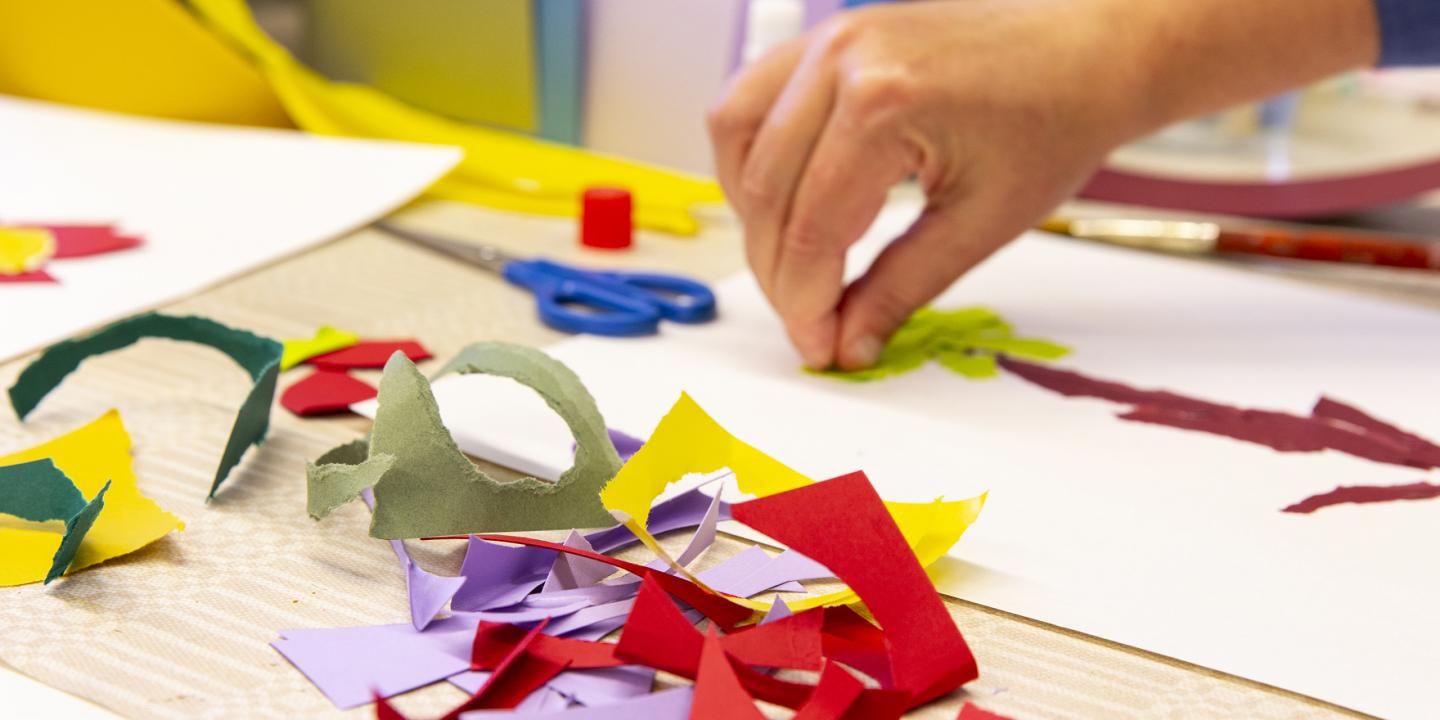Radošā darbnīca bērniem: roka līmē krāsaina papīra formas uz balta lapas, uz galda redzamas šķēres, līme un izgriezti papīra gabali.