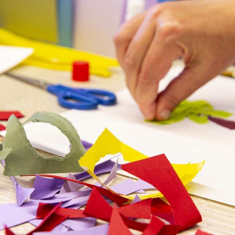 Radošā darbnīca bērniem: roka līmē krāsaina papīra formas uz balta lapas, uz galda redzamas šķēres, līme un izgriezti papīra gabali.