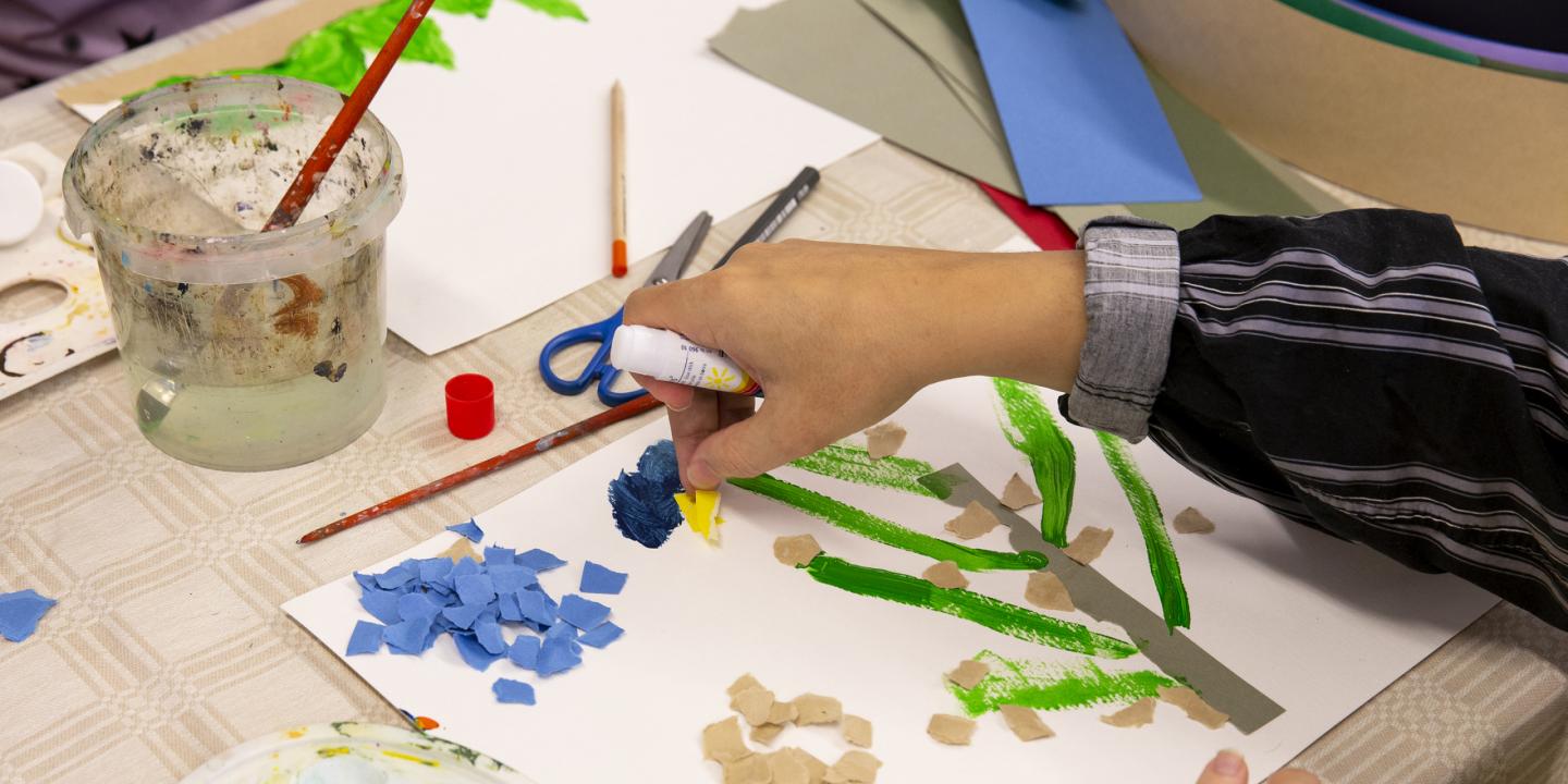 Radošā darbnīca bērniem: roka ar līmes zīmuli piestiprina krāsaina papīra gabaliņus uz zīmējuma ar zaļām krāsas līnijām; uz galda redzamas šķēres, otas, ūdens trauks un papīra izgriezumi.