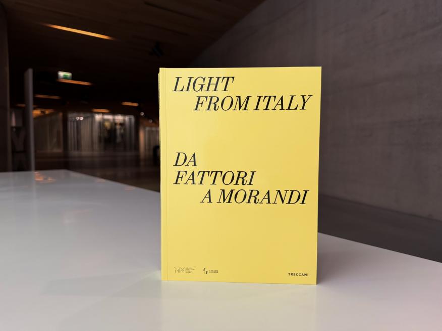 Uz balta galda izstādīta dzeltena grāmata ar melnu uzrakstu “Light from Italy. Da Fattori a Morandi”; fonā redzama muzeja telpa ar tumšām sienām un apgaismojumu.