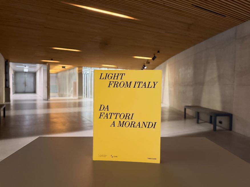 Dzeltena grāmata “Light from Italy. Da Fattori a Morandi” novietota uz galda modernā muzeja interjerā ar koka griestiem un plašu gaiteni fonā.