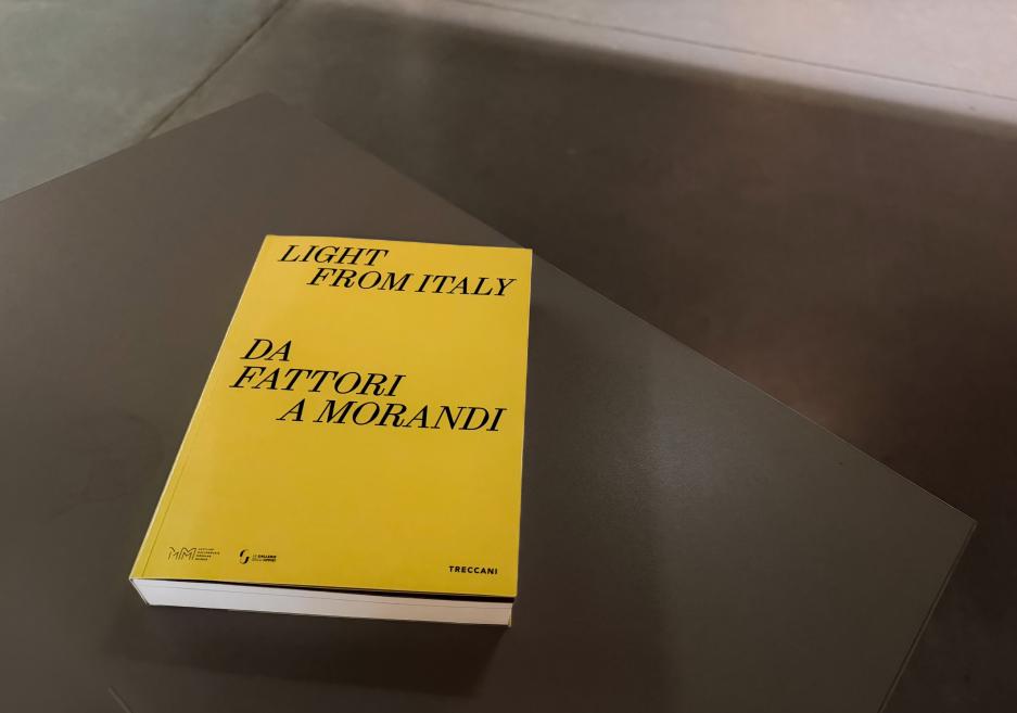Dzeltena grāmata “Light from Italy. Da Fattori a Morandi” guļ uz tumšas galda virsmas, fotografēta no augšas ar vieglu perspektīvu un ēnu fonā.