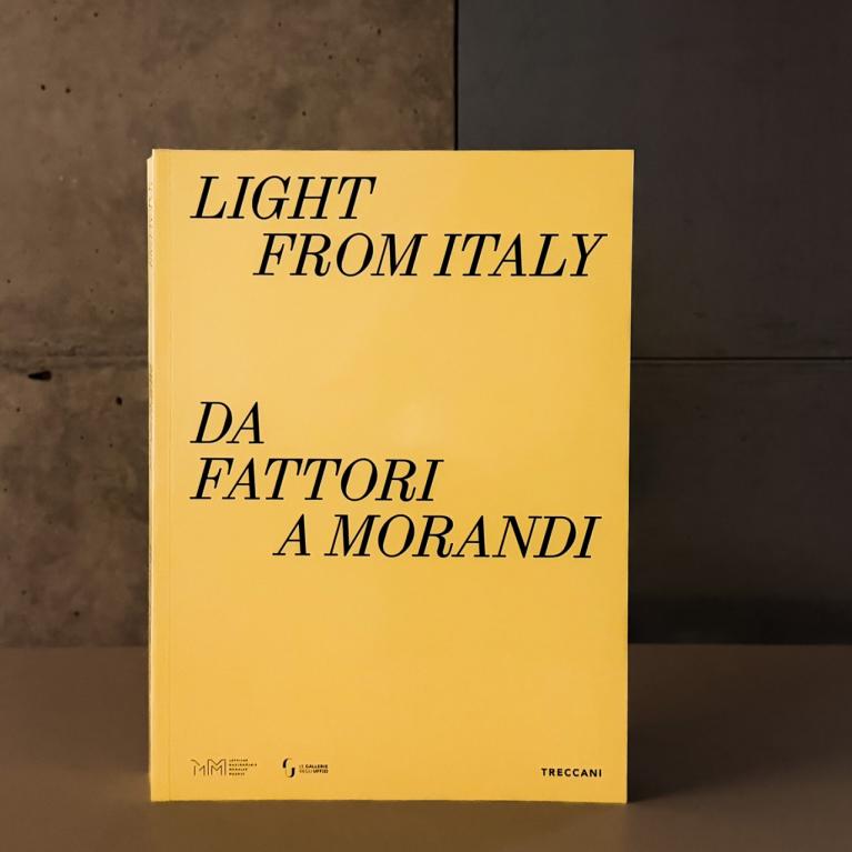 Dzeltenas krāsas grāmatas vāks ar melnu uzrakstu “Light from Italy. Da Fattori a Morandi”, novietots uz galda mūsdienīgā muzeja telpā; fonā redzama betona siena un kāpnes.