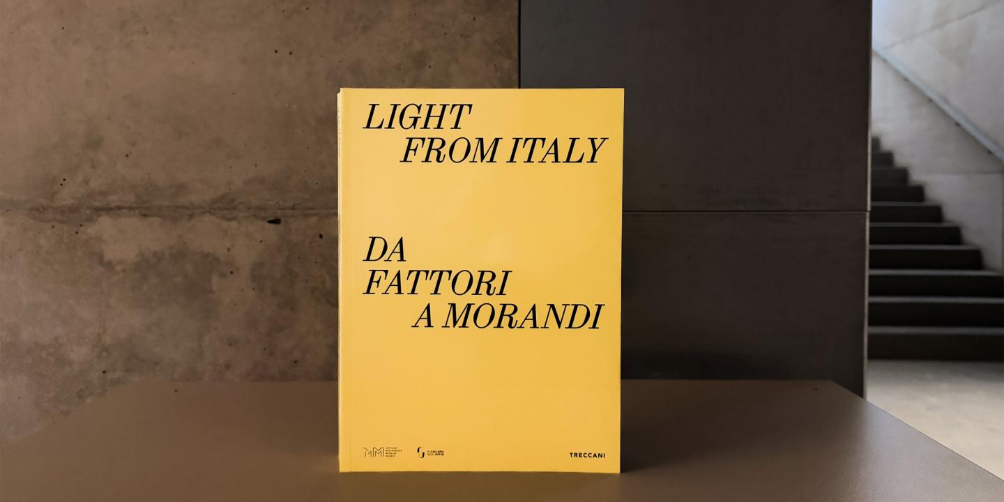 Dzeltenas krāsas grāmatas vāks ar melnu uzrakstu “Light from Italy. Da Fattori a Morandi”, novietots uz galda mūsdienīgā muzeja telpā; fonā redzama betona siena un kāpnes.