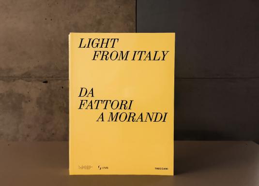 Dzeltenas krāsas grāmatas vāks ar melnu uzrakstu “Light from Italy. Da Fattori a Morandi”, novietots uz galda mūsdienīgā muzeja telpā; fonā redzama betona siena un kāpnes.