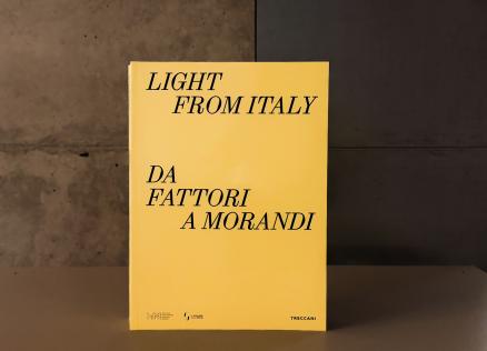 Dzeltenas krāsas grāmatas vāks ar melnu uzrakstu “Light from Italy. Da Fattori a Morandi”, novietots uz galda mūsdienīgā muzeja telpā; fonā redzama betona siena un kāpnes.