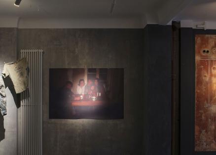 Instalācija izstāžu telpā: uz sienas pie auklas piespraustas nodilušas drēbes, centrā fotogrāfija ar cilvēkiem pie galda vājā apgaismojumā, bet labajā pusē liela jauktas tehnikas darba virsma ar tekstu un nelielu foto, industriālā, pelēkā vidē.