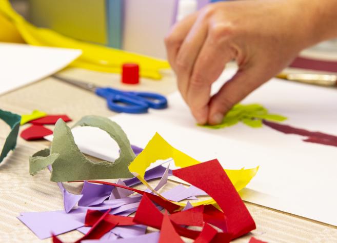 Radošā darbnīca bērniem: roka līmē krāsaina papīra formas uz balta lapas, uz galda redzamas šķēres, līme un izgriezti papīra gabali.